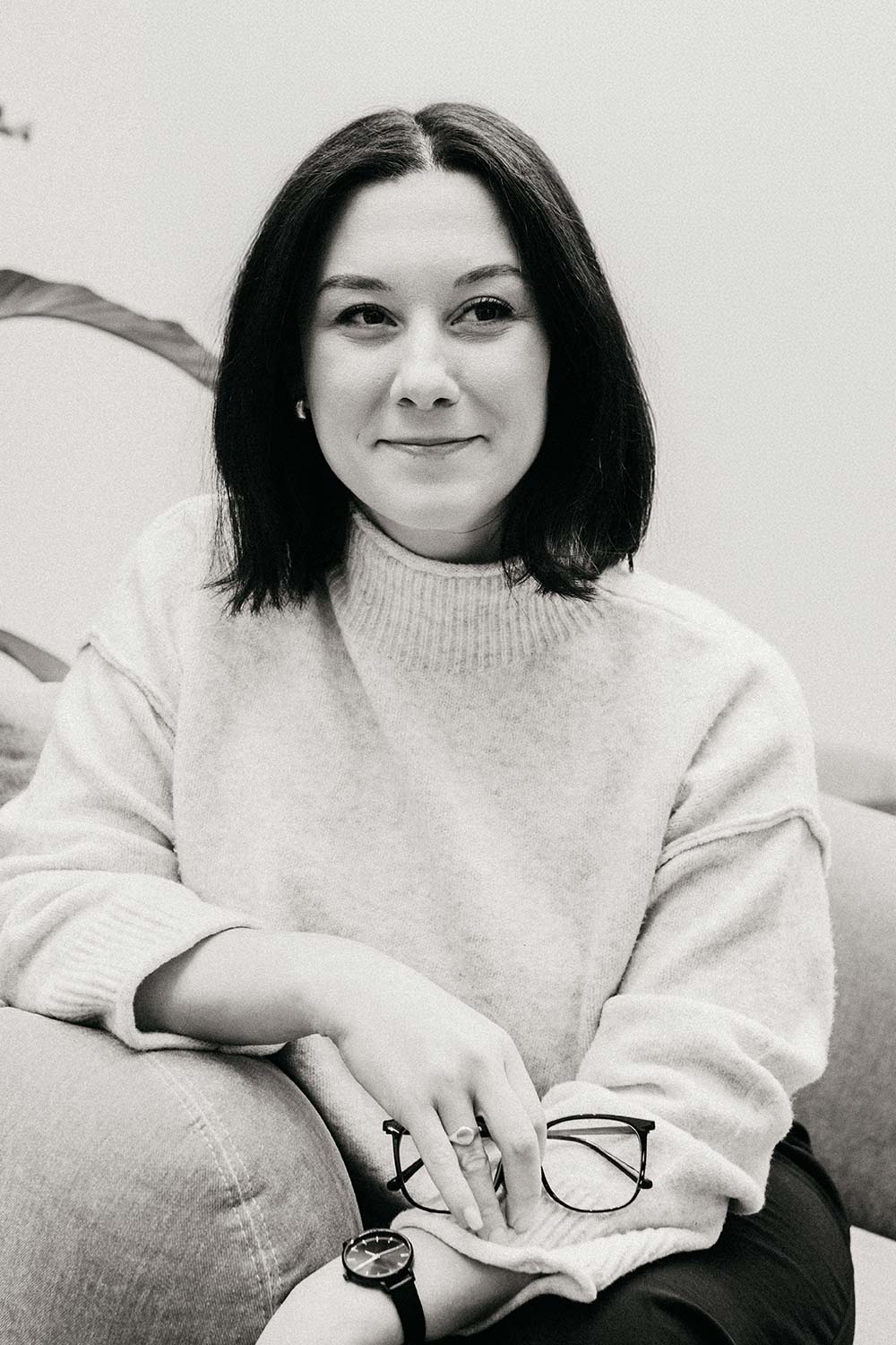 Woman with short dark hair in a light knit sweater sits on a couch, smiling slightly, holding glasses in her hand and wearing a wristwatch.