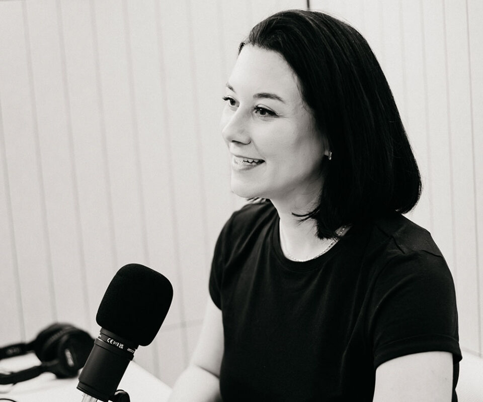 Woman with short dark hair speaking into a tabletop microphone in a studio setting.