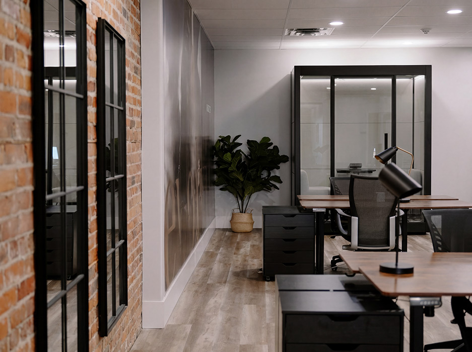 Open-plan office with an exposed brick wall on the left, desks, filing cabinets, a potted plant, and a glass-enclosed meeting room in the back.