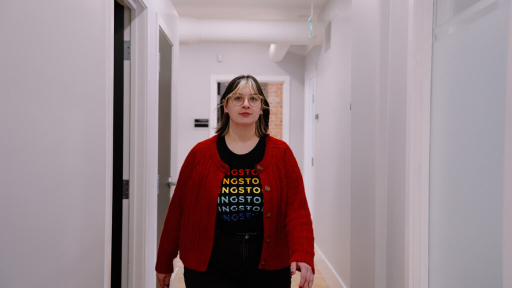 Person walking down a white hallway wearing a bright red cardigan over a black shirt with multicolored text, facing the camera.