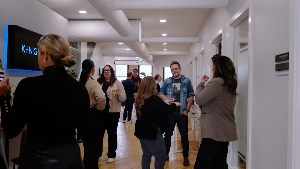 Coworkers mingling and chatting in a bright office hallway with drinks in hand during a networking event.