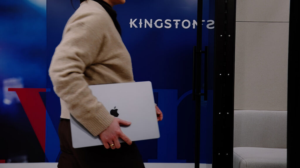 Person walking past a blue wall while carrying a closed silver laptop under their arm