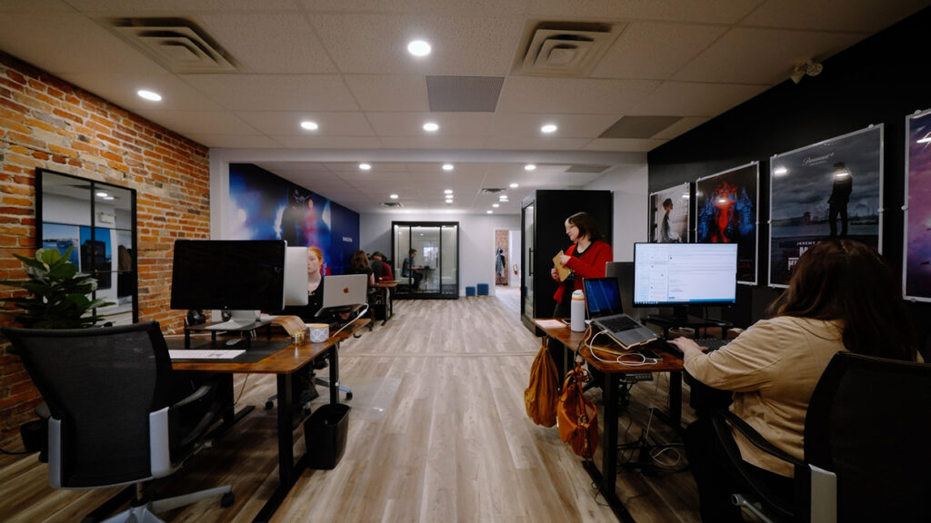 Open-plan office with a brick wall on the left, desks and laptops, posters on the right, and a woman in red standing near a desk.