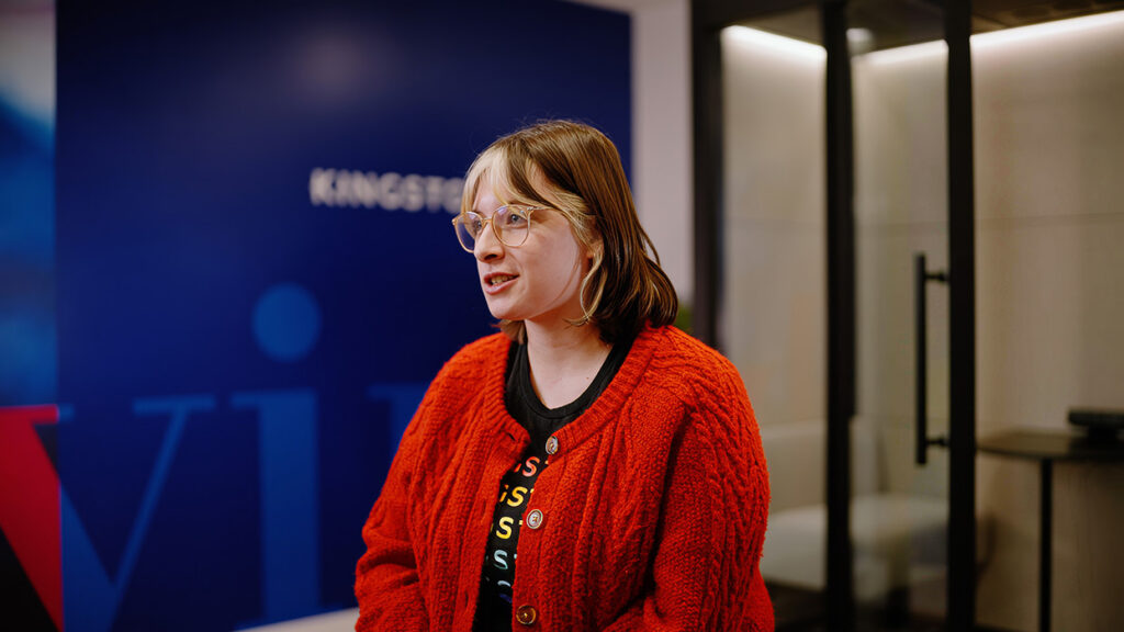 Woman with short blonde hair and glasses wearing a bright red cardigan, speaking in a modern office lobby.