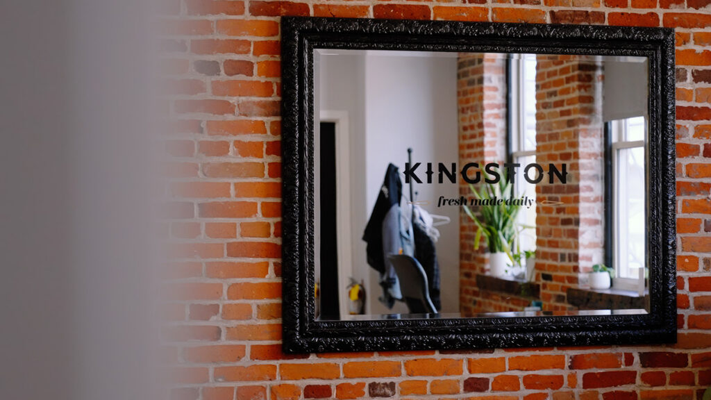 Ornate black-framed mirror on a brick wall reflecting a clothing rack and a plant; Kingston—fresh made daily is written on the glass.