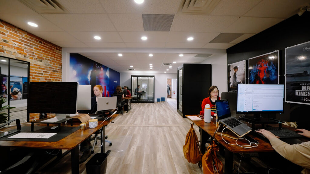 Open coworking space with multiple desks and people working on laptops, brick accent wall on the left and poster wall on the right.