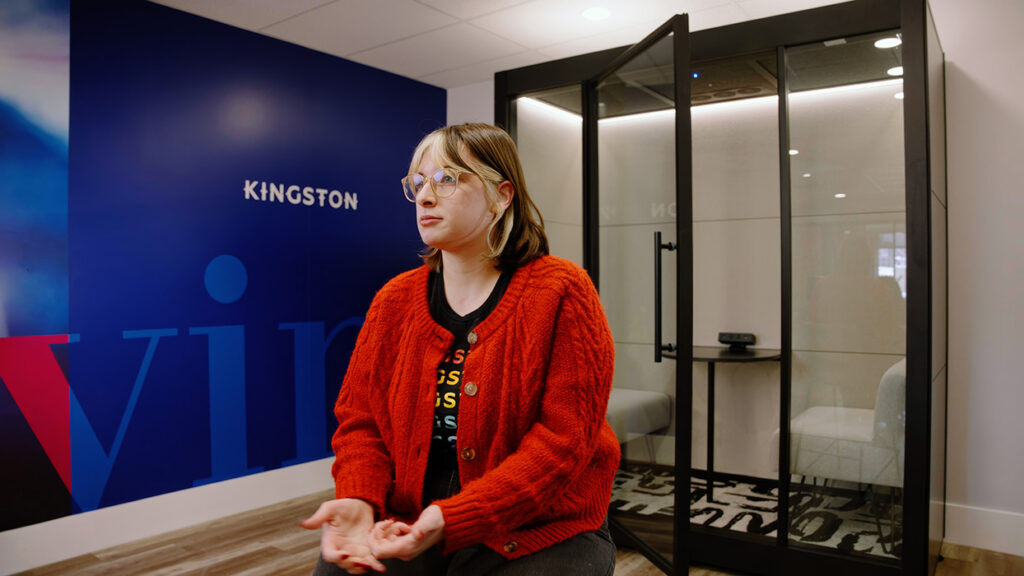 Woman with a red cardigan sits in a modern office lobby, Kingston branding on a blue wall behind her.