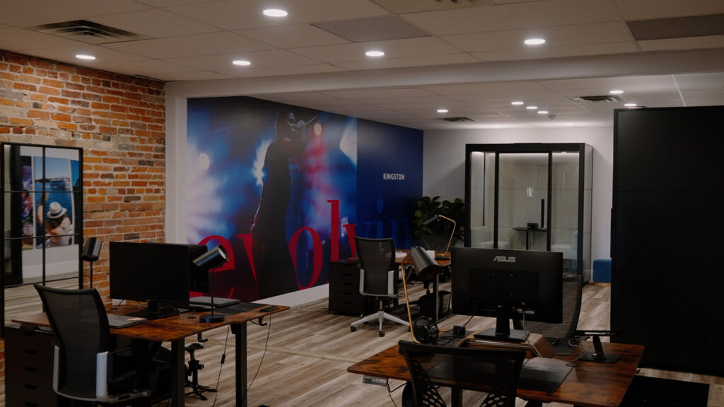 Open-plan office with wooden desks, computer monitors, and desk lamps, a brick accent wall on the left, and a large stage-like mural in the back.