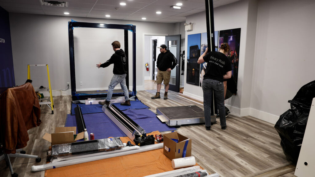 Room under setup with a large black metal frame on a blue mat; three people in casual clothing work on a project, with tools, boxes, and foam on the floor.
