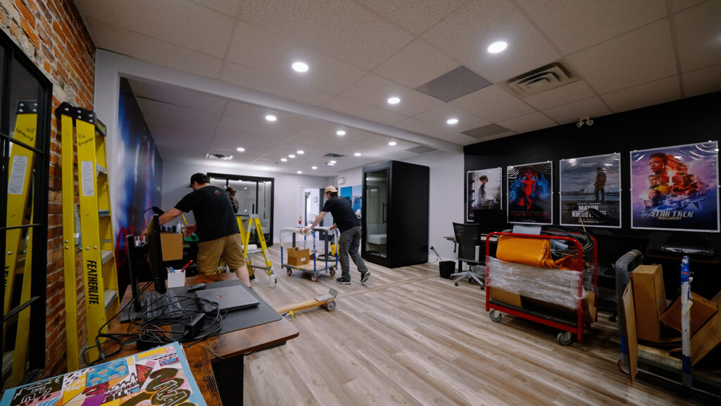Two workers set up a workspace in a studio/office with ladders, carts, and posters on a black wall, under bright ceiling lights