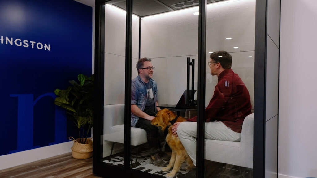 Two men inside a glass meeting pod chat, with a golden retriever seated between them on a white couch.