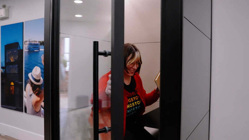 Smiling woman in a red cardigan sits inside a glass-enclosed booth, giving a thumbs up.
