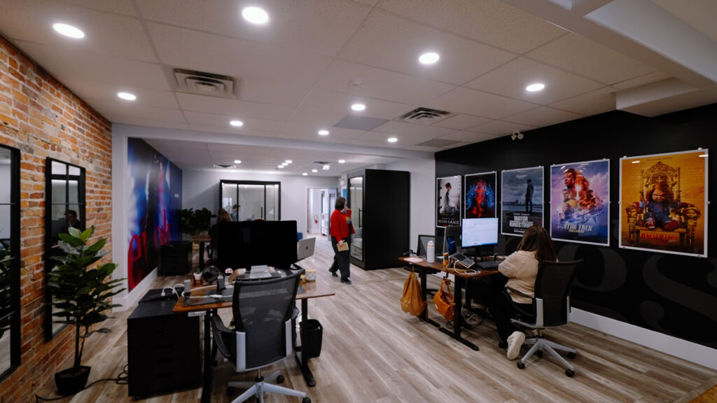 Open office space with a brick wall, desks, and computer monitors; posters line a black wall as a person in a red jacket walks in the background.