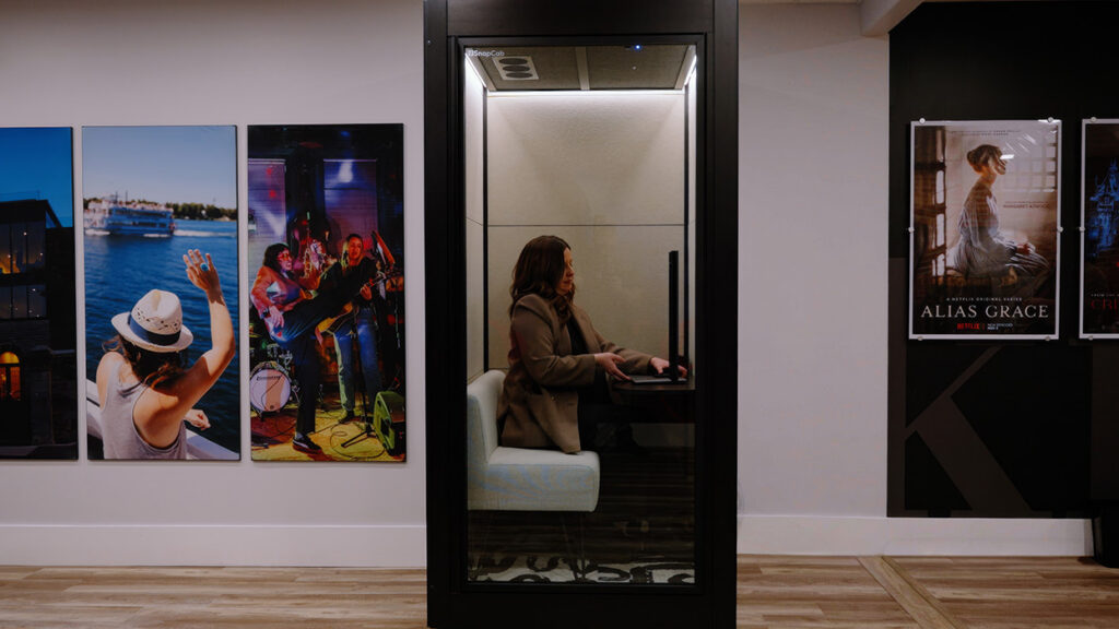 Woman in a private glass booth working on a laptop amid large wall posters in a gallery hallway.
