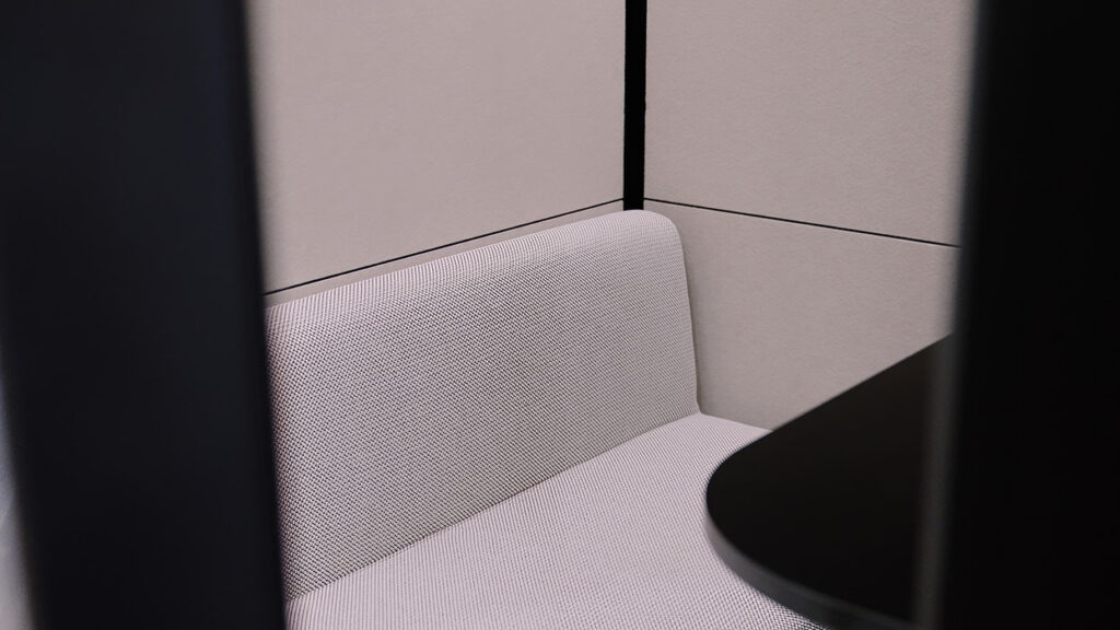 Corner view of a light gray fabric loveseat in a cubicle with a round black table nearby.