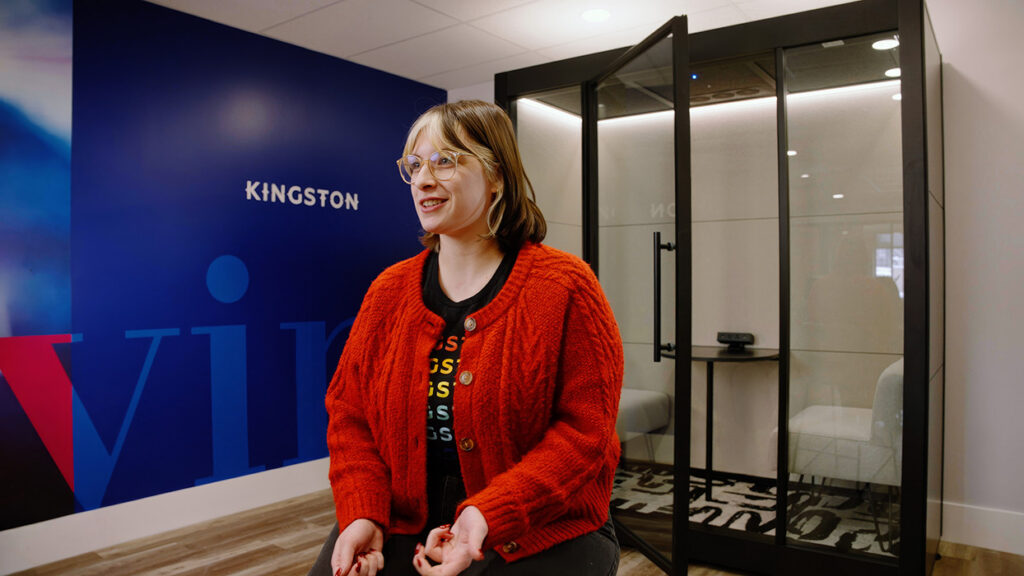 Smiling woman with glasses in a bright orange cardigan sits in a modern office lobby with Kingston signage on a blue wall behind her.