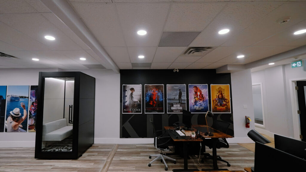 Office lobby with a dark accent wall displaying five movie posters, a glass-walled privacy pod on the left, and a wooden desk with chairs in the center-right.