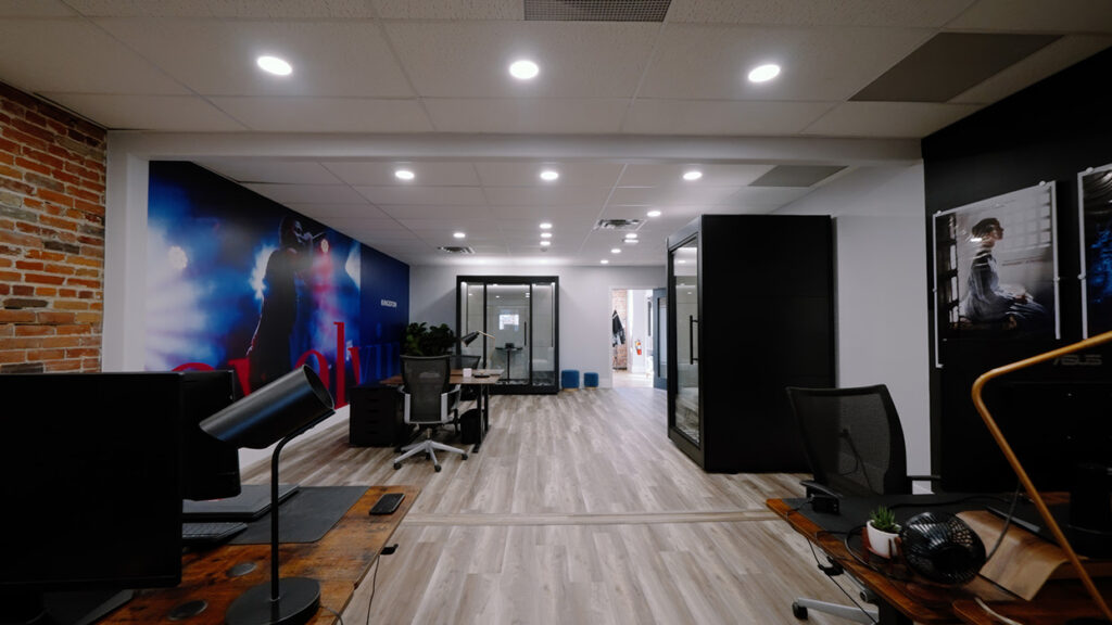 Open-plan office with wooden floors, exposed brick wall, a large mural on the left, desks and chairs, and glass-walled rooms in the back.