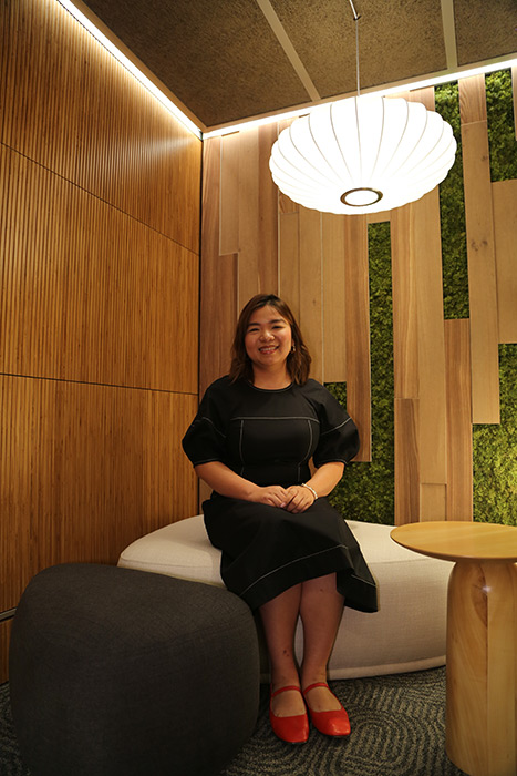 Smiling woman in a black dress sits on a white curved sofa in a wood-paneled lounge with green wall panels and a large white circular pendant light.