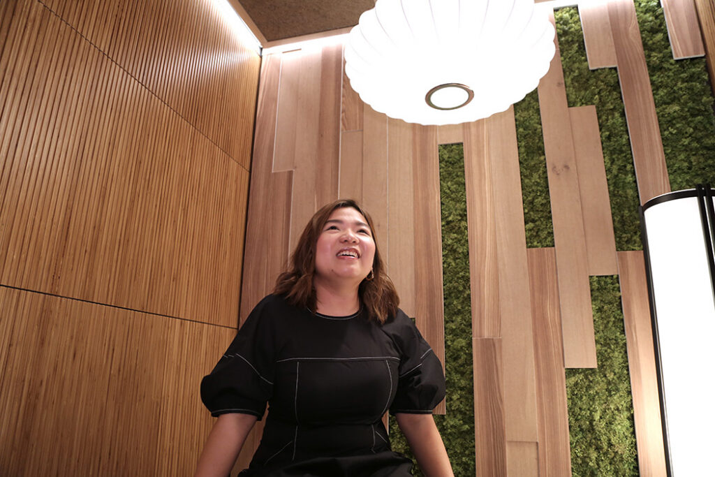 Smiling woman seated in a wood-paneled room with moss walls and a large white pendant light overhead.