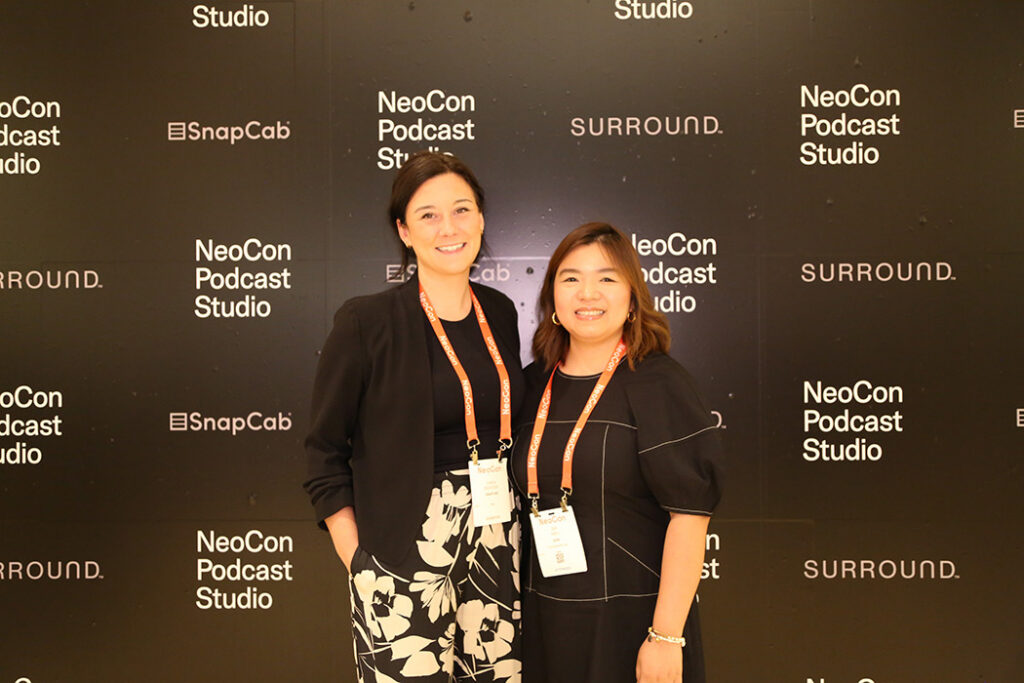 Two women posing together in front of a NeoCon Podcast Studio backdrop at a conference, smiling and wearing event lanyards.