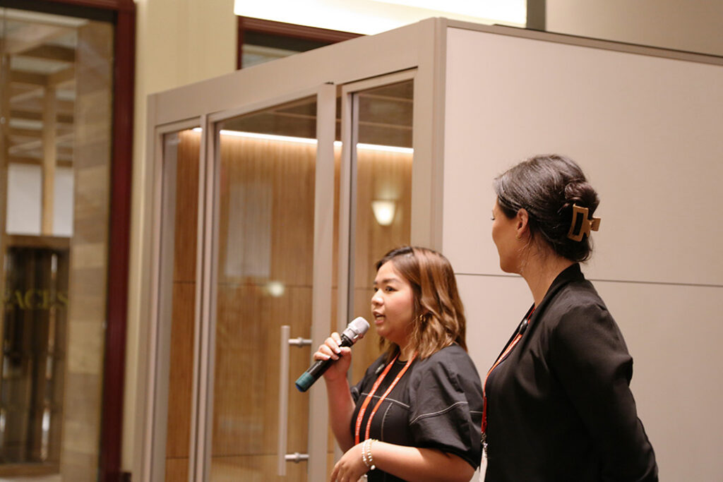 Two women at a conference; one speaks into a microphone while the other stands nearby indoors.