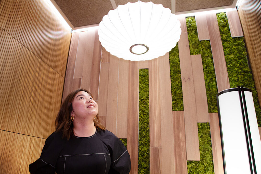 Woman in a black top stands in a wood-paneled room with green moss wall panels, looking up at a large circular white ceiling light.