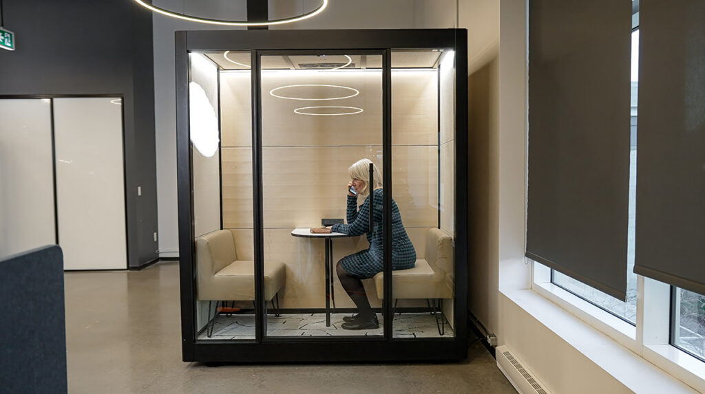 A woman sits at a small round table inside a glass-walled phone booth, talking on a cellphone.