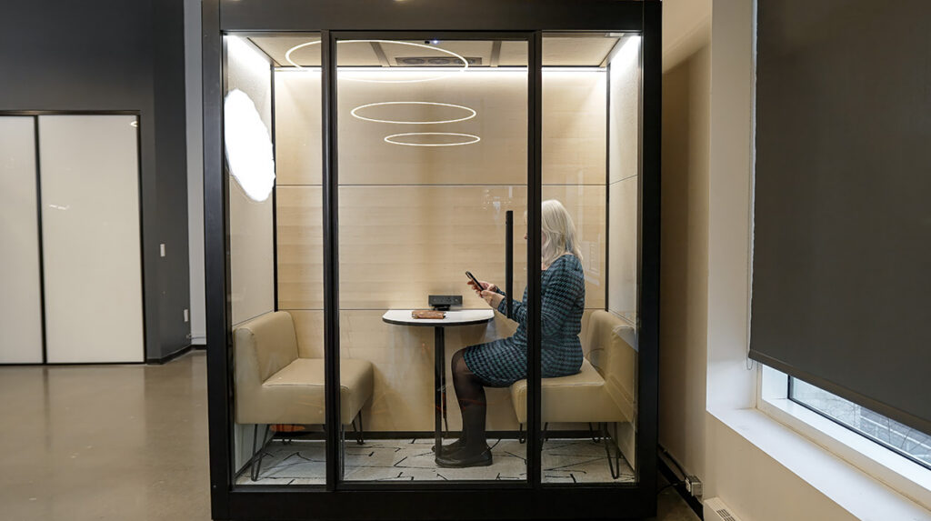 Woman with blond hair seated in a glass-enclosed office booth, using a smartphone at a small round table.
