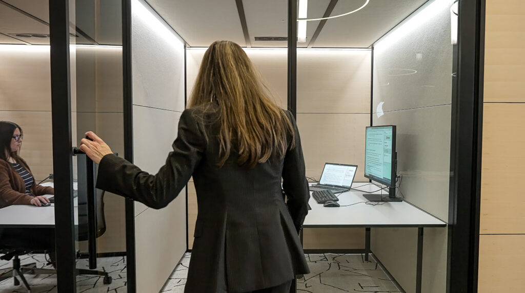 Businesswoman in a dark blazer entering a glass-walled private office with a computer on the desk inside.