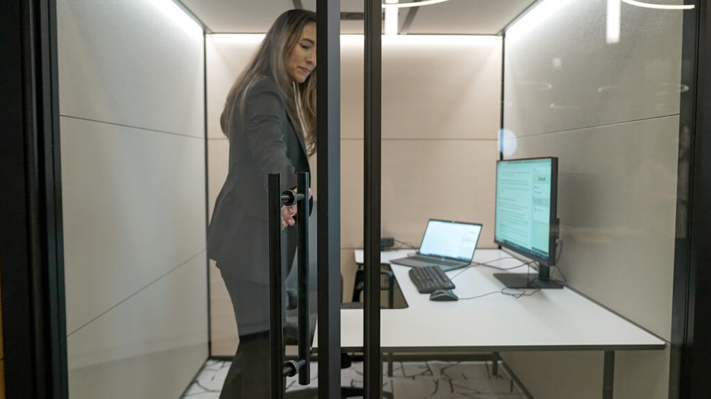 Woman in a dark blazer stands inside a glass privacy pod, reaching for the door handle beside a white desk with a laptop and monitor.]','