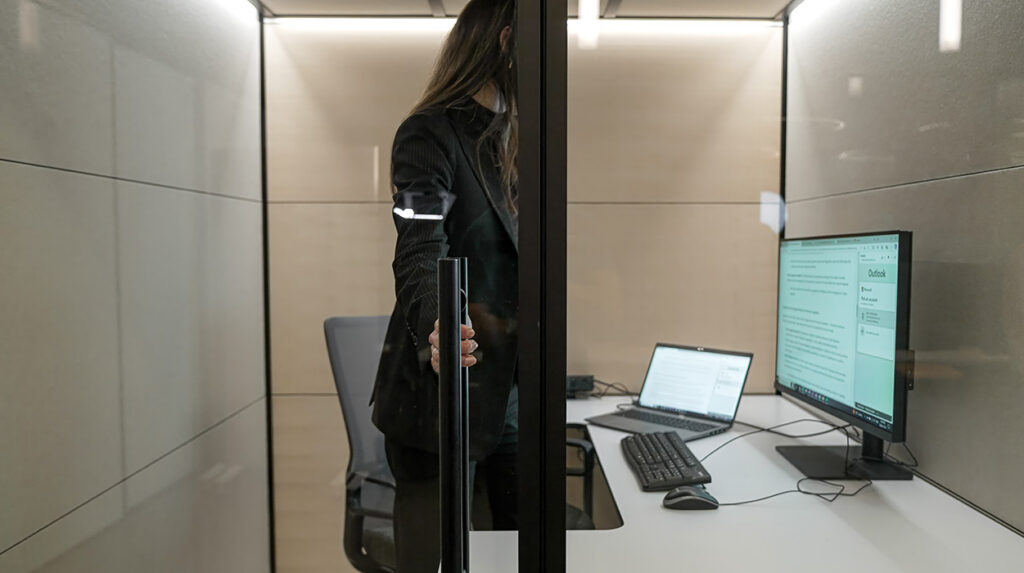 Person stands in a glass-walled office cubicle, hand on the door handle, with a laptop and monitor showing Outlook on the desk.