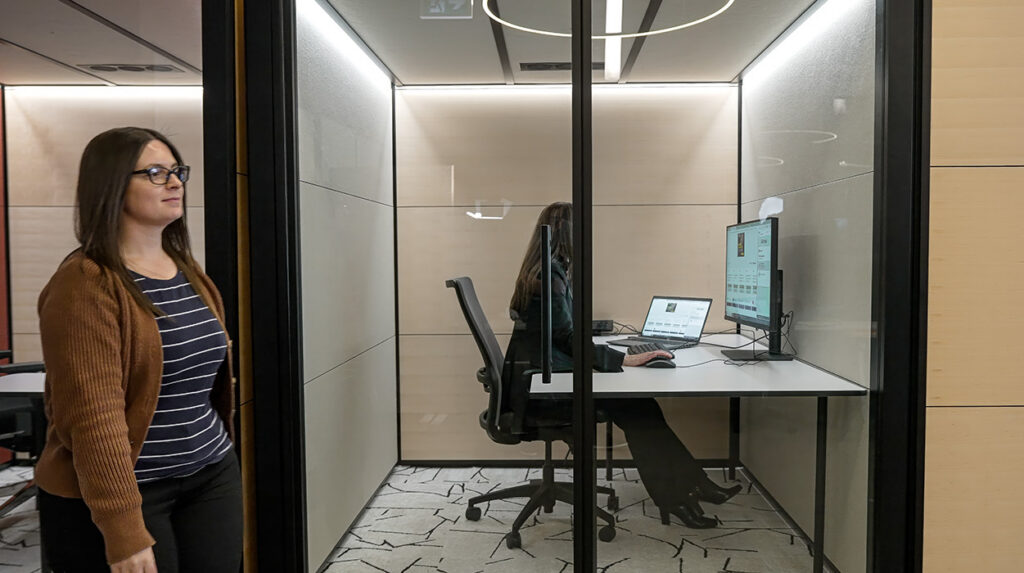 A woman with glasses walking past a glass-walled office booth while another person sits at a computer inside the booth.