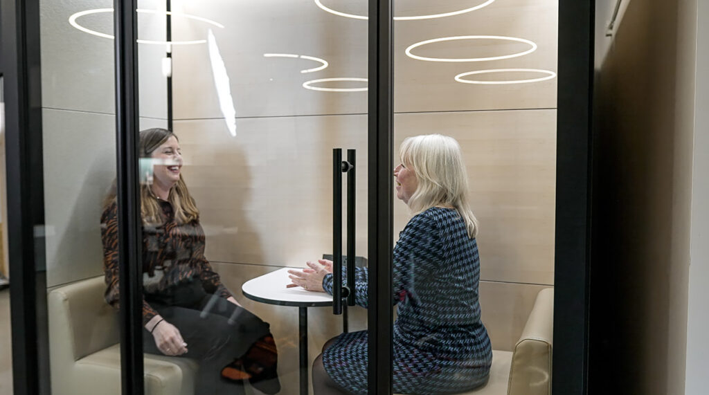 Two women sit in a glass-walled meeting pod, smiling and talking across a small round table.