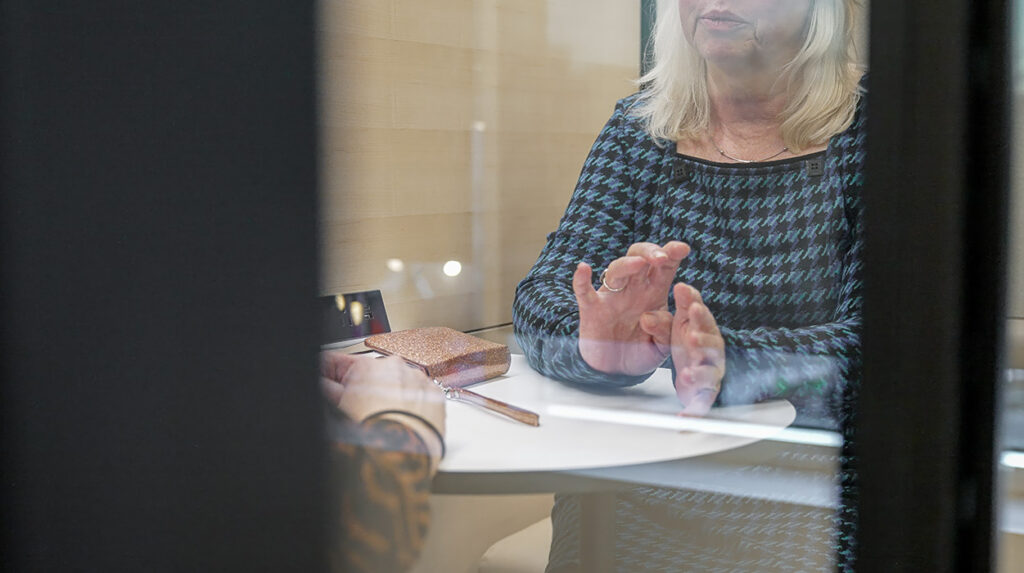 Woman in a blue patterned top behind a glass partition, gesturing with her hands over a white table with a glittery clutch nearby.