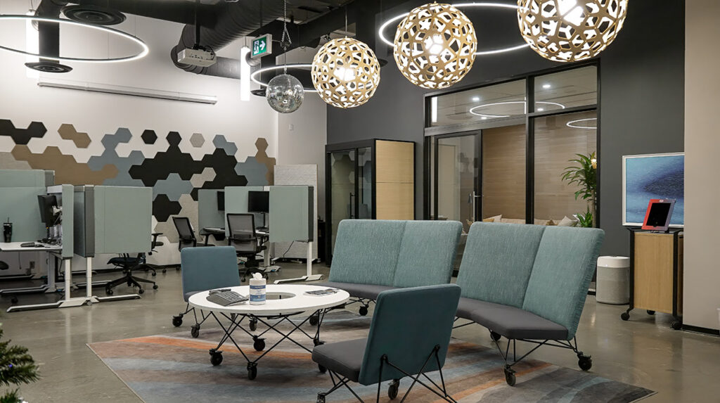 Open-plan office lounge with teal chairs, a white circular table on wheels, and a high hexagon-patterned wall behind workstations under large pendant lights.
