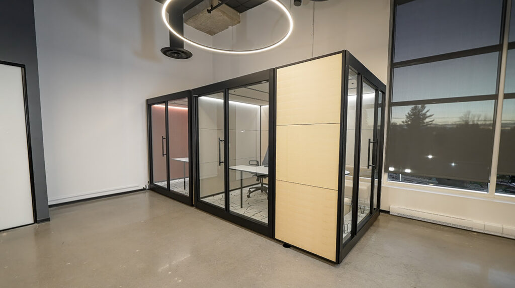 Modern office with glass-walled private pods, a circular ceiling light, and large windows; beige panels separate a row of glass offices on a concrete floor.