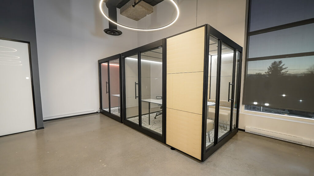 Modern office with glass-walled meeting pods and a circular pendant light above the space, along a wall with a large window nearby.