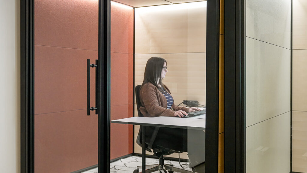 Woman with glasses sits at a white desk in a glass-enclosed office pod, typing on a keyboard.