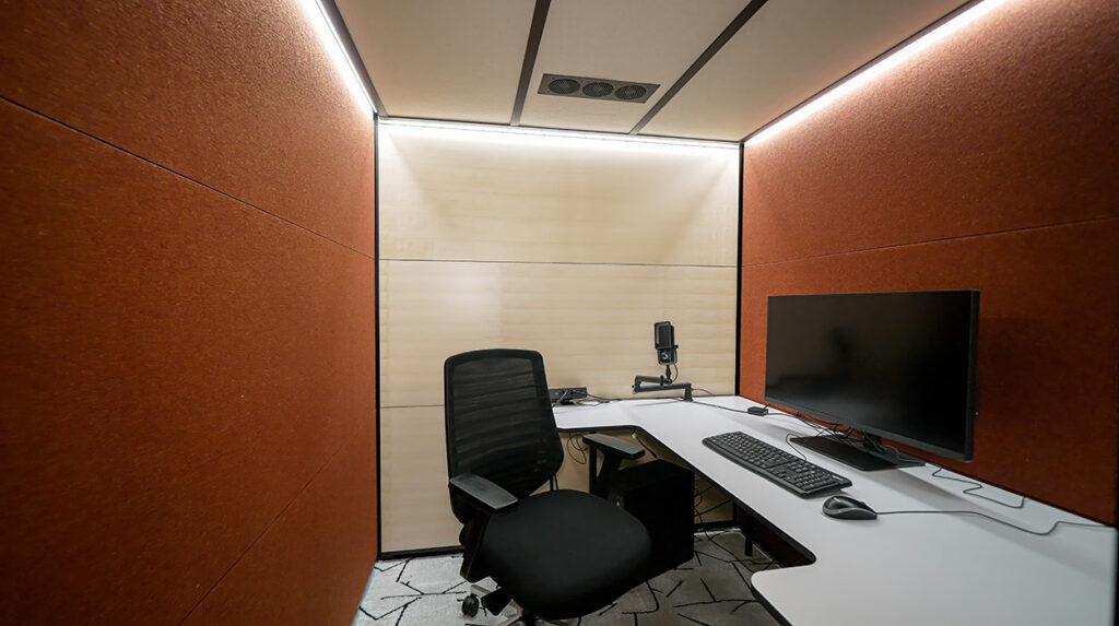 Soundproof recording booth with orange-brown acoustic panels and a white desk setup, including a monitor, keyboard, mouse, and microphone on a stand in the corner.