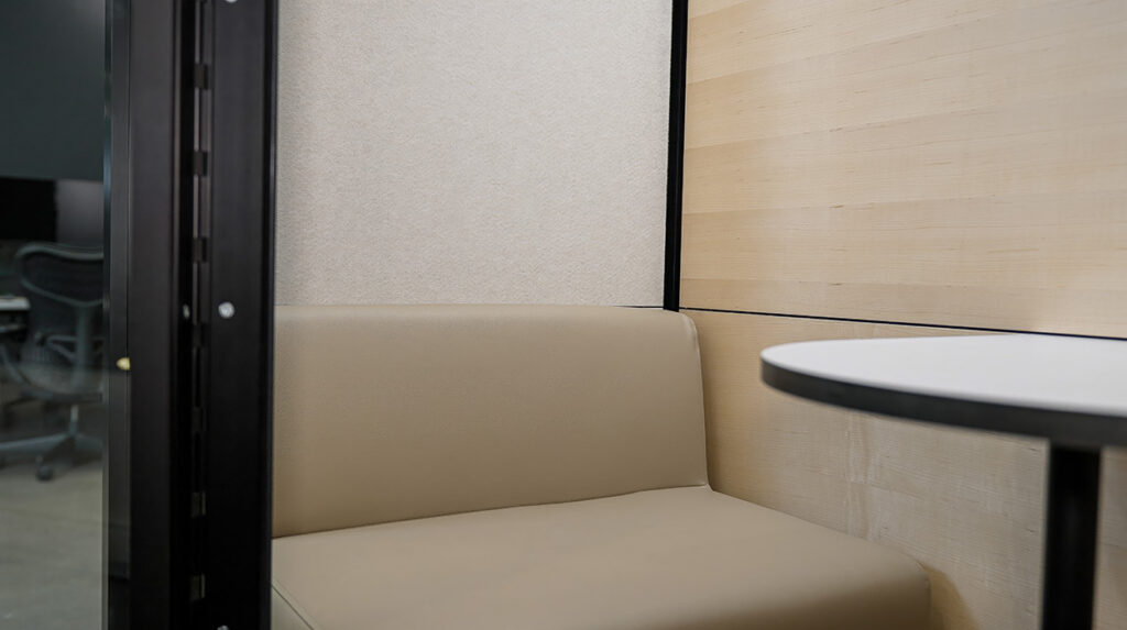 Beige upholstered booth in a corner with a round black-edged table and light wood wall panels; glass door to the left hints at an office area beyond.