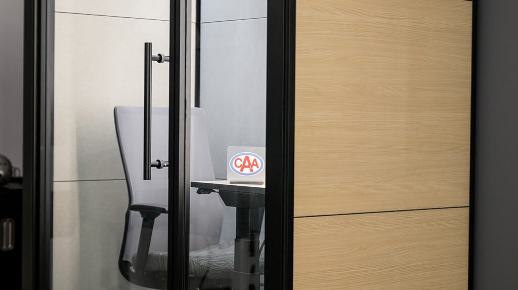 Glass-walled meeting room with a grey office chair and a desk, featuring a CAA logo on a small sign.