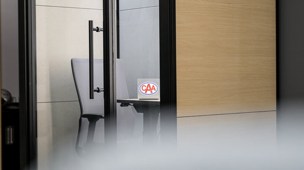 View into a small conference room through glass doors: a gray chair and table with a circular logo placard on the desk inside, frosted glass at the bottom edge.