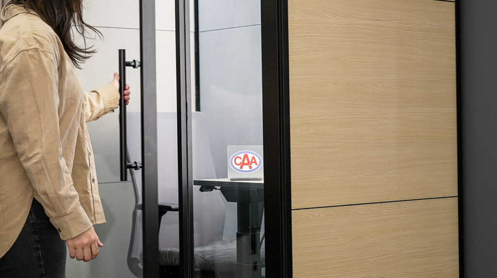 Person in a beige jacket opens a glass door into a private office or meeting area; a CAA sticker is visible on the table inside.