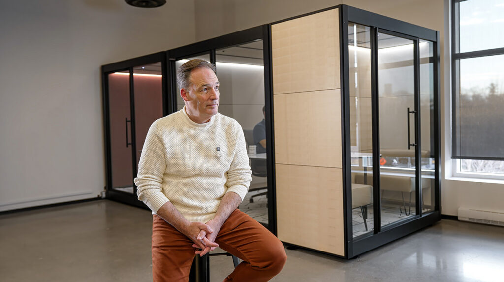 Man seated in a cream knit sweater in a modern office, with glass-walled meeting pods behind him.