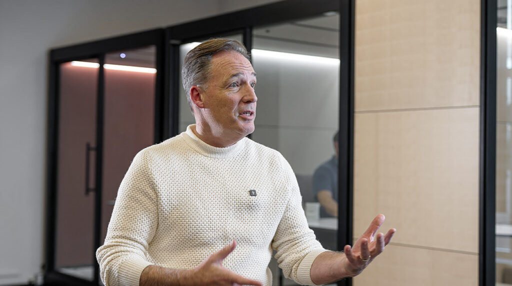 Middle-aged man in a cream knit sweater speaks with animated hand gestures in a modern office setting
