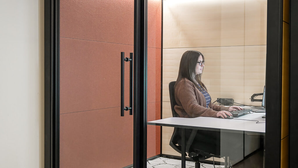 Woman wearing glasses, brown cardigan, and striped shirt working at a desk inside a glass-walled office, typing on a keyboard