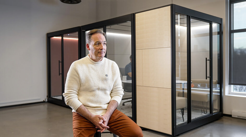 Man sitting on a stool in a modern office, cream sweater and orange pants, with glass-walled meeting pods behind him.