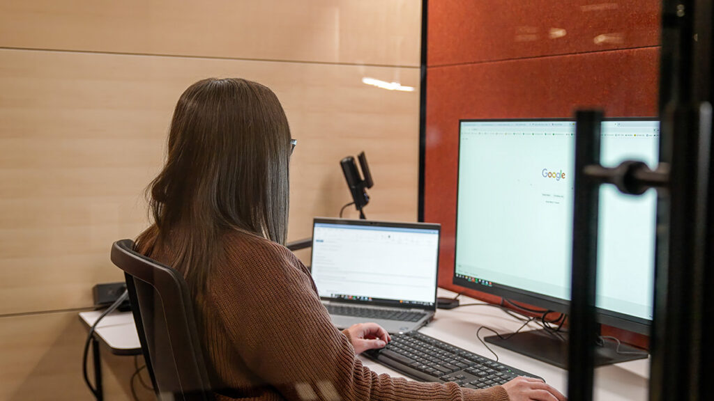 Person seated at a desk, typing on a laptop with a second monitor showing Google in a modern office setting