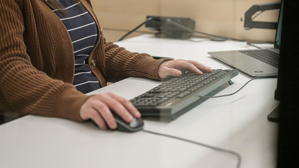 Person typing on a keyboard with a mouse on a white desk, laptop nearby in a work setup